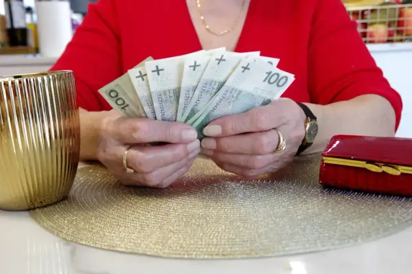 Dłonie trzymające banknoty 100 zł. Zastanawiasz się, komu przysługuje przeliczenie emerytury? Obok złoty wazon i czerwona szkatułka.