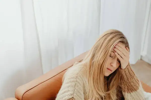 Kobieta z blond włosami, w swetrze, siedzi na kanapie, trzymając się za czoło. Wygląda na zmęczoną, jakby miała l4 na wypowiedzeniu.
