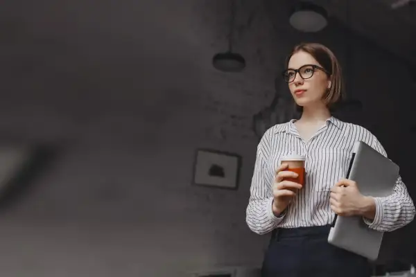 Kobieta w okularach trzymająca kawę i laptopa.