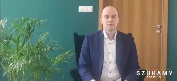 Mężczyzna w garniturze siedzi w fotelu obok rośliny, na tle zielonej ściany i drewnianych drzwi.