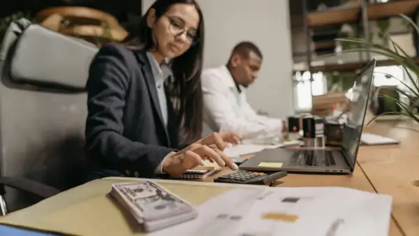 Kobieta w okularach liczy na kalkulatorze, obok stos banknotów i laptop.