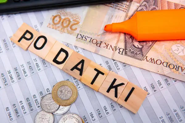 Drewniane klocki z napisem "PODATKI", banknot 200 zł, monety i pomarańczowy marker na arkuszu kalkulacyjnym. Myśląc o przyszłości, warto pamiętać o podatku katastralnym.