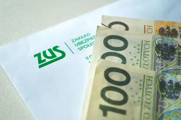 ZUS i polskie banknoty 100 zł. Pieniądze leżą obok koperty z logo ZUS, symbolizując finanse i ubezpieczenia społeczne.
