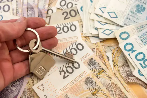 Dłoń trzyma klucz z breloczkiem w kształcie domu na tle polskich banknotów. Obniżka stopy procentowej może oznaczać tańsze kredyty hipoteczne.