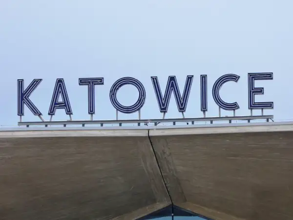 Napis "KATOWICE" z niebieskimi neonami na tle jasnego nieba, nad betonową konstrukcją.